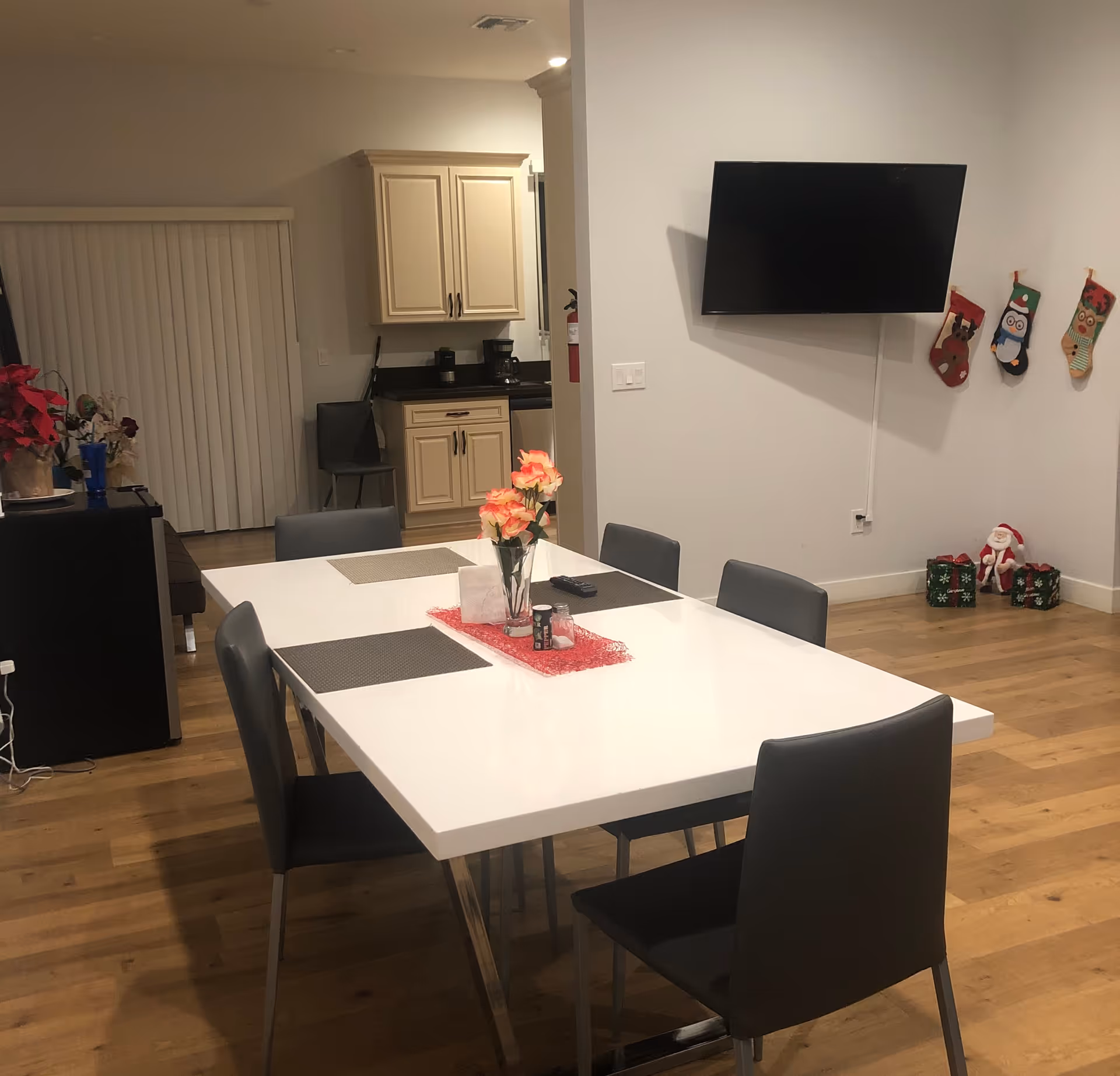 Interior view of a dining area with a white rectangular table surrounded by six gray chairs. The table has a vase with flowers and placemats. In the background, there is a kitchen area with cream-colored cabinets and a coffee maker. On the right wall, a flat-screen TV is mounted above four Christmas stockings and holiday decorations on the floor. The floor is wooden, and there is a sliding door with vertical blinds on the left side.