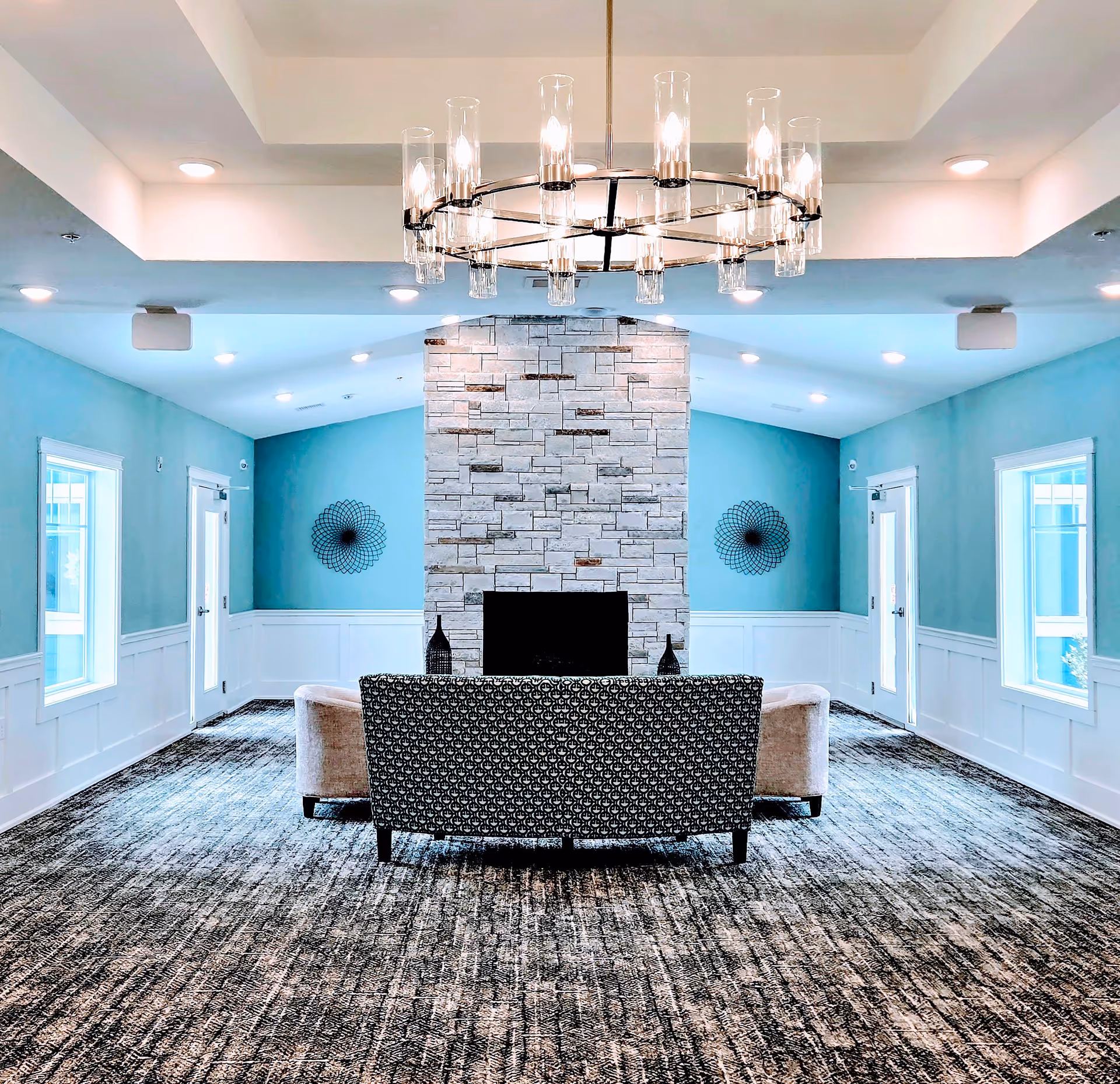 A spacious and modern common area with a large stone fireplace centered on the far wall. In front of the fireplace are two beige armchairs and a patterned sofa. The walls are painted light blue with white wainscoting, and there are two windows and two doors on either side of the fireplace. A large, circular chandelier with multiple lights hangs from the ceiling. The floor is covered with a textured carpet in shades of gray.