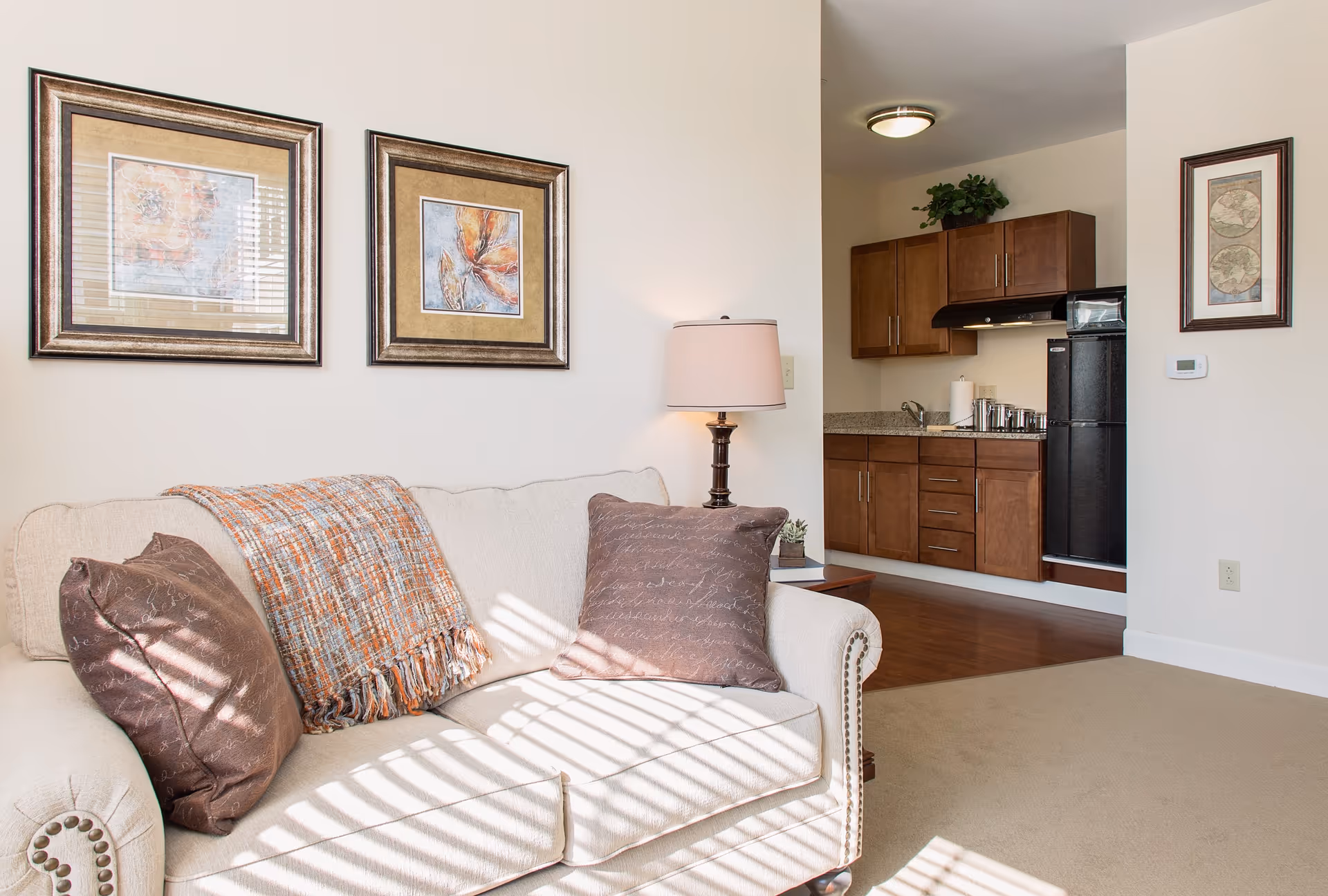 A cozy living room area with a beige sofa adorned with two brown pillows and a multicolored throw blanket. Above the sofa are two framed floral artworks. To the right, there is a wooden side table with a lamp and a small plant. In the background, a small kitchen area with wooden cabinets, a black refrigerator, a sink, and a stove hood is visible. The walls are light-colored, and sunlight streams in through a window, casting shadows on the sofa.