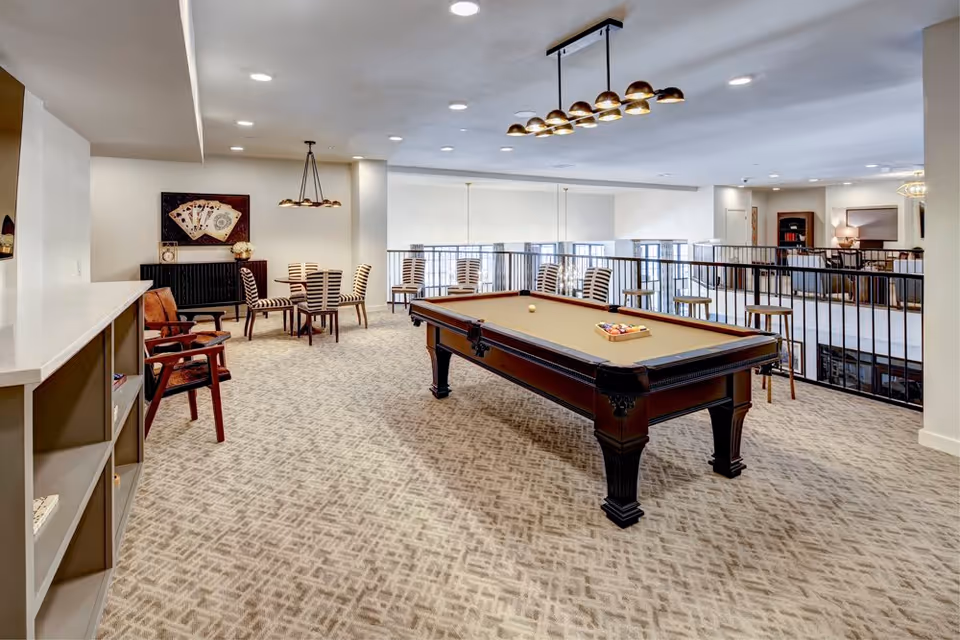 A spacious recreational room with a pool table in the center, surrounded by several chairs and small tables. The room has patterned carpet flooring, modern ceiling lights, and large windows with blinds. There is a railing overlooking a lower level, and the decor includes a wall art piece featuring playing cards.