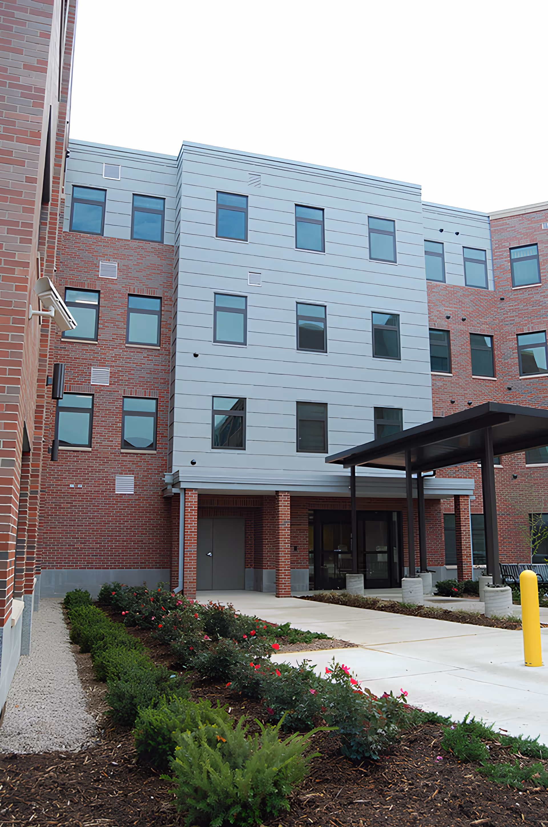 Front entrance of a multi-story brick and metal-clad building with a covered drop-off and landscaped walkway.