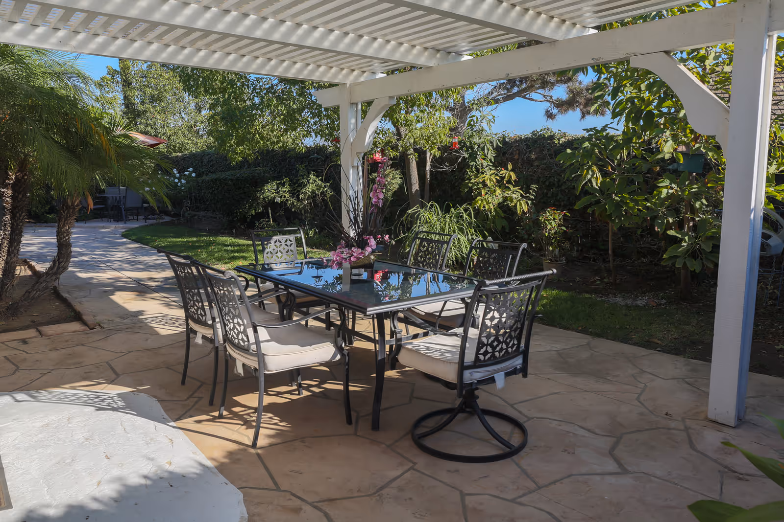 Shaded outdoor patio with a glass-top dining table and six metal chairs under a white pergola, surrounded by garden plants and trees.