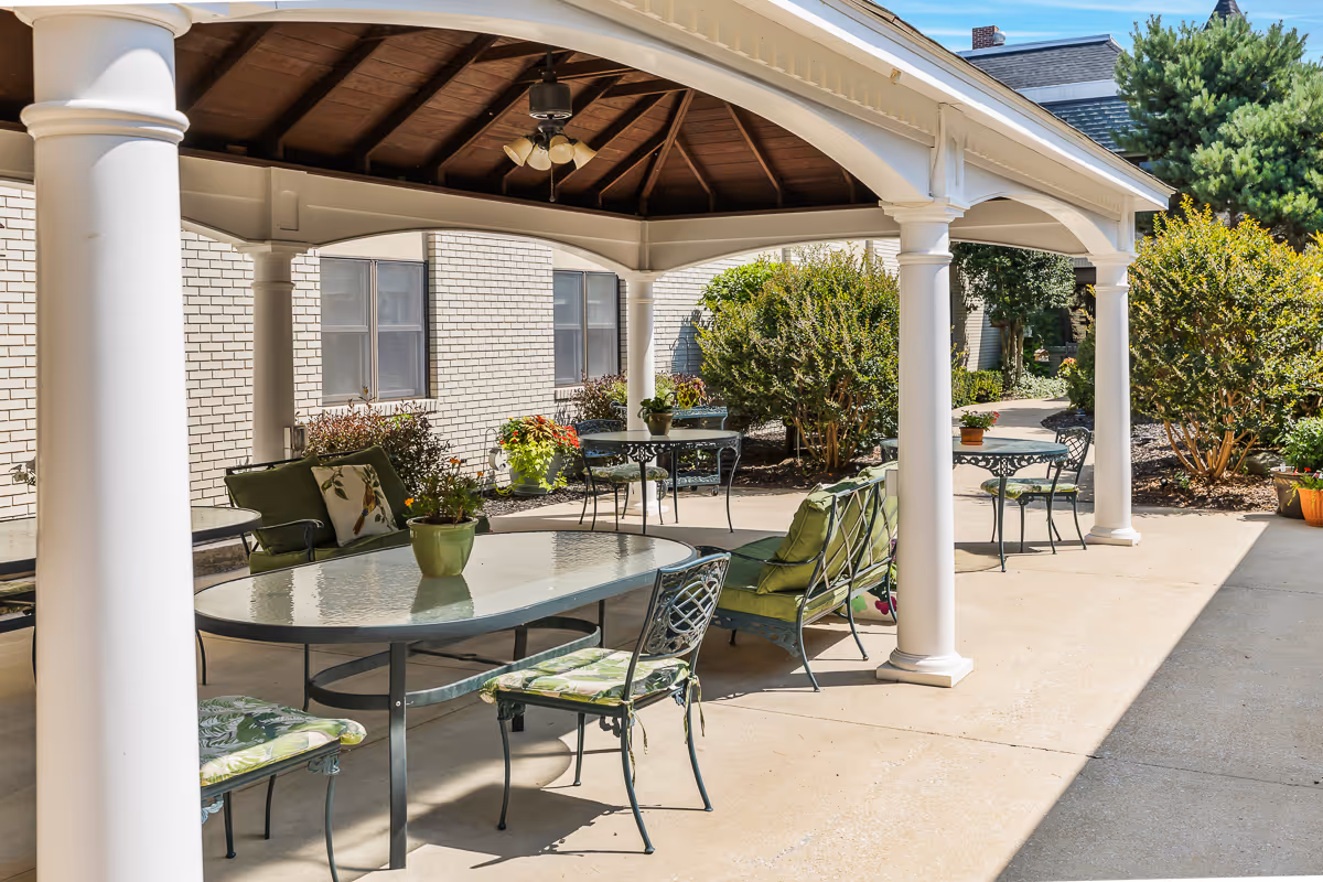 Outdoor covered patio area with white columns and a wooden ceiling, furnished with glass-top tables and metal chairs with green cushions. Surrounding the patio are bushes and potted plants, with a white brick building in the background under a clear blue sky.