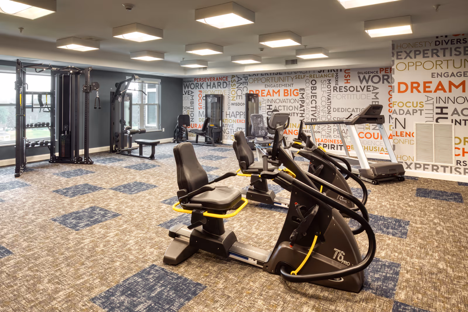 A fitness room in a senior living facility with exercise equipment including recumbent bikes, a treadmill, and a multi-station weight machine. The room has large windows letting in natural light and a wall decorated with motivational words like 'Dream Big', 'Focus', and 'Opportunity'. The floor is carpeted with a patterned design.