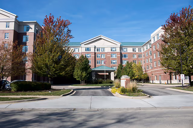 Front exterior view of a large multi-story senior living facility building with a green roof, brick facade, and several trees with autumn foliage in front. A driveway leads to the entrance with a covered drop-off area.