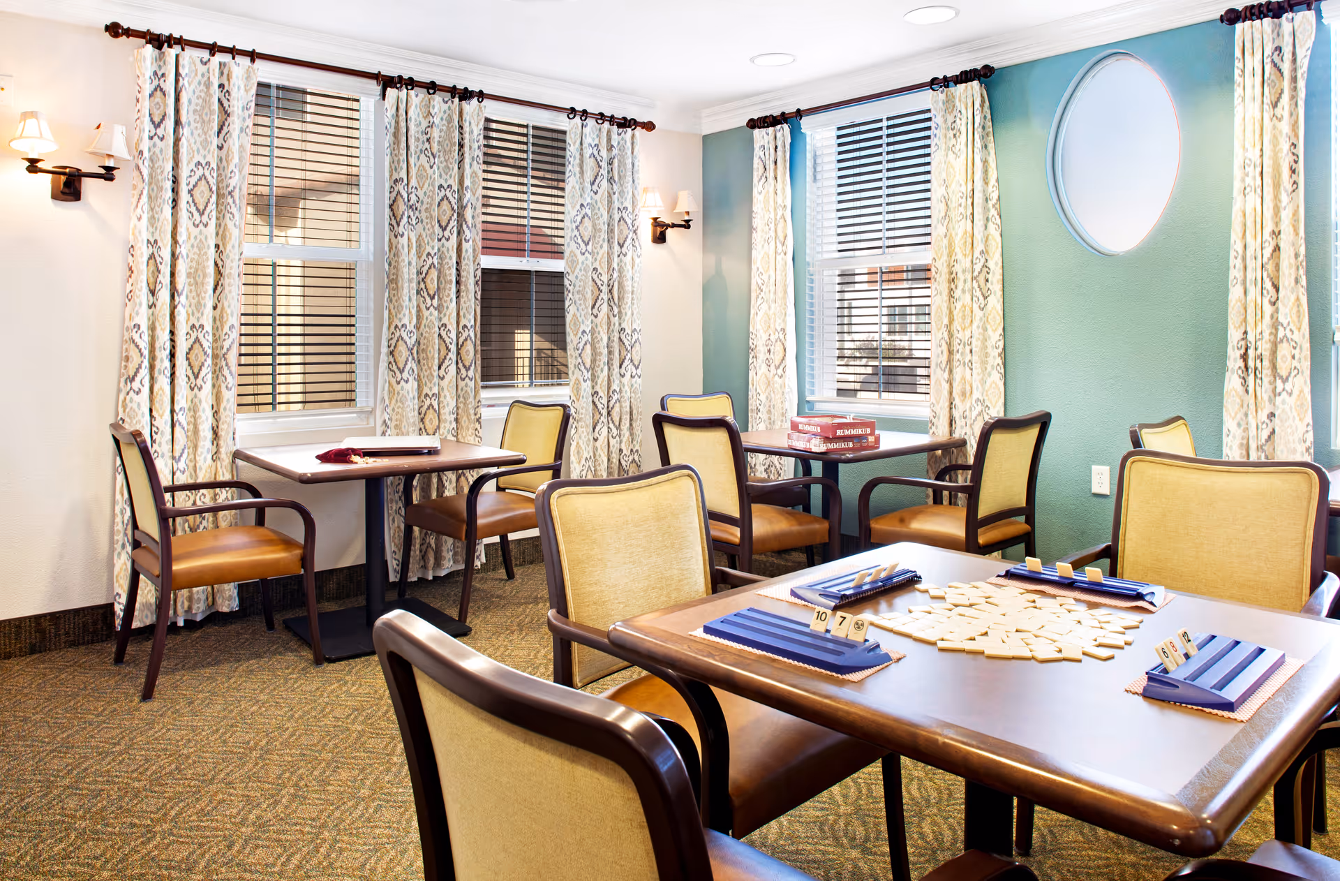 A well-lit room with multiple tables and chairs arranged for playing board games. One table in the foreground has a Scrabble game set up with tiles and racks. The room has patterned curtains on the windows, wall sconces with lights, and a circular window on a green accent wall.
