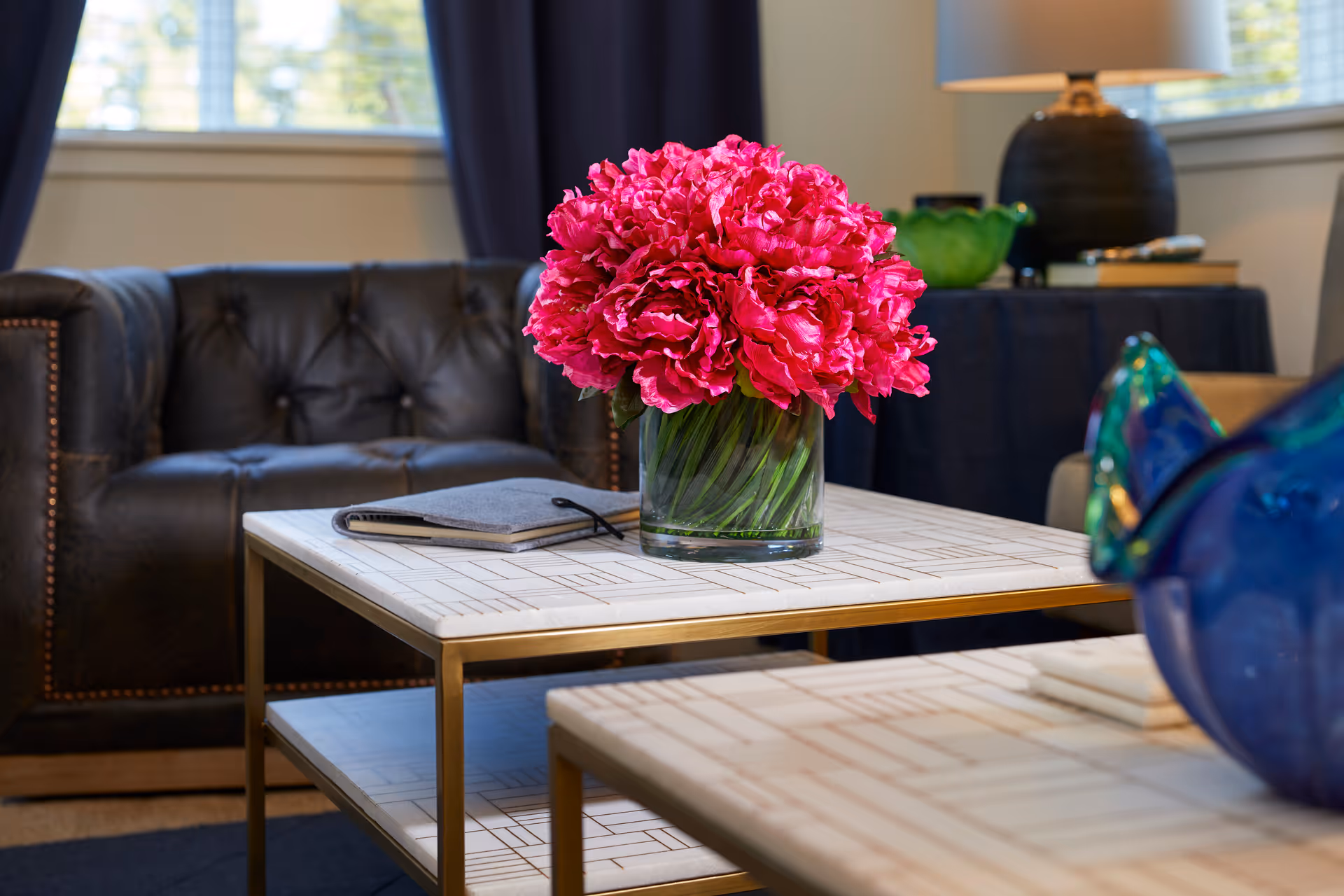 A close-up view of a living room area featuring a glass vase with bright pink flowers on a white patterned coffee table with gold legs. In the background, there is a dark leather tufted armchair, a table with a dark tablecloth, a lamp, and windows with curtains.