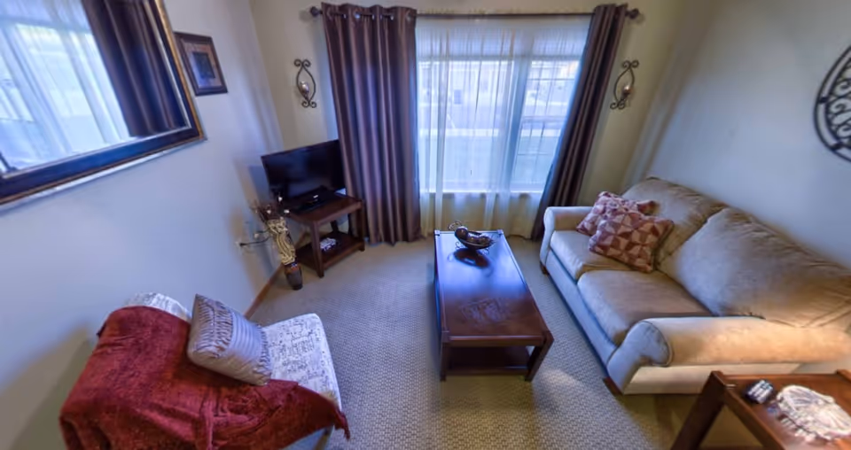 Cozy living room with a sofa, armchair, coffee table, TV on a stand, and a large curtained window.