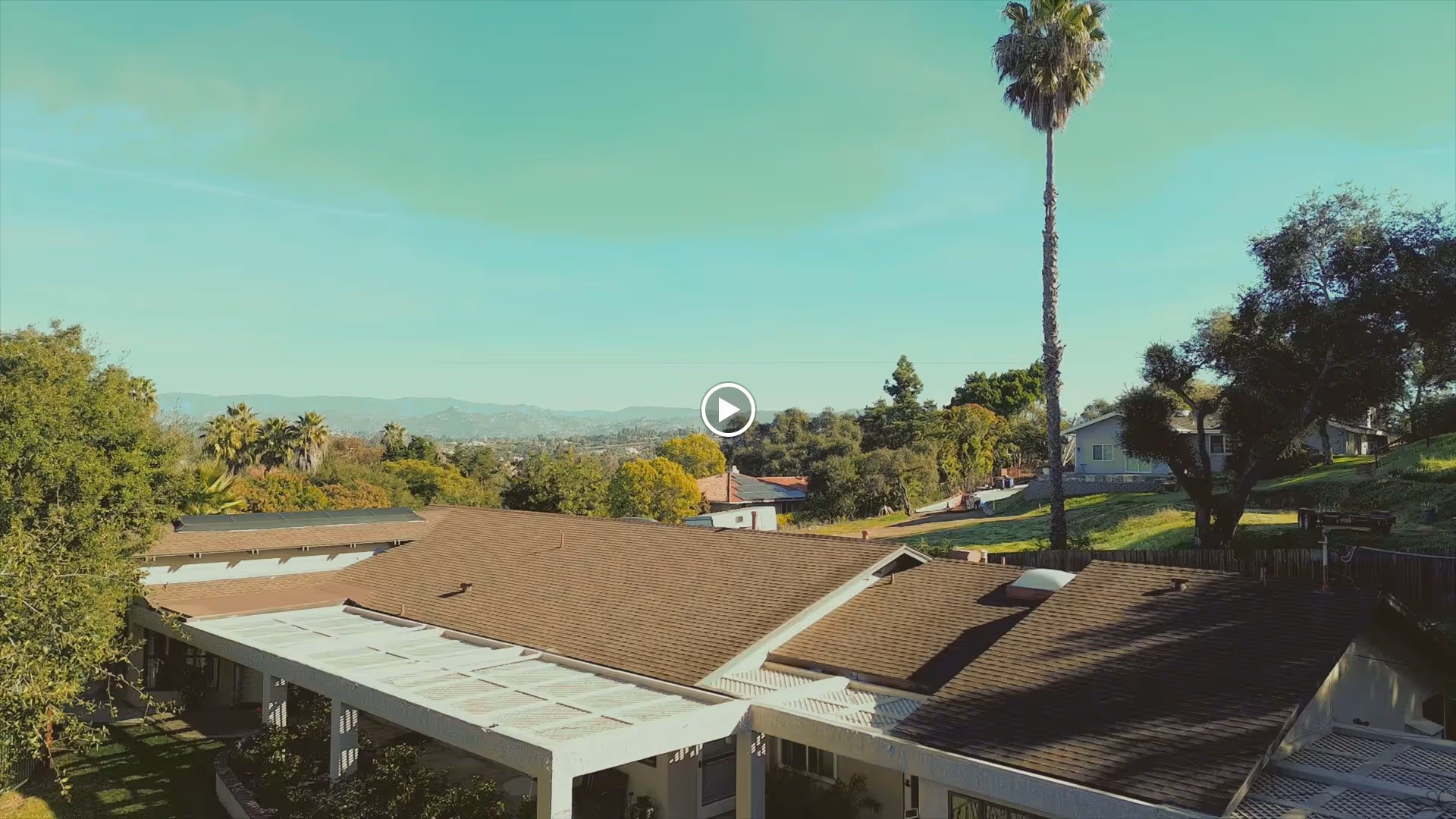 Aerial view of a single-story assisted living facility with brown shingle roofs, surrounded by trees and greenery under a clear blue sky. A tall palm tree and other trees are visible on the property, with distant hills in the background.