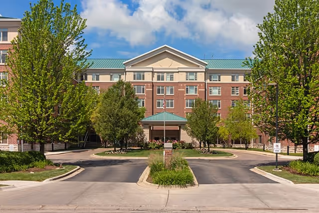 Front exterior view of a multi-story senior living facility building with a green roof, brick facade, and a covered entrance. The driveway is circular with landscaped greenery and trees on both sides under a partly cloudy sky.