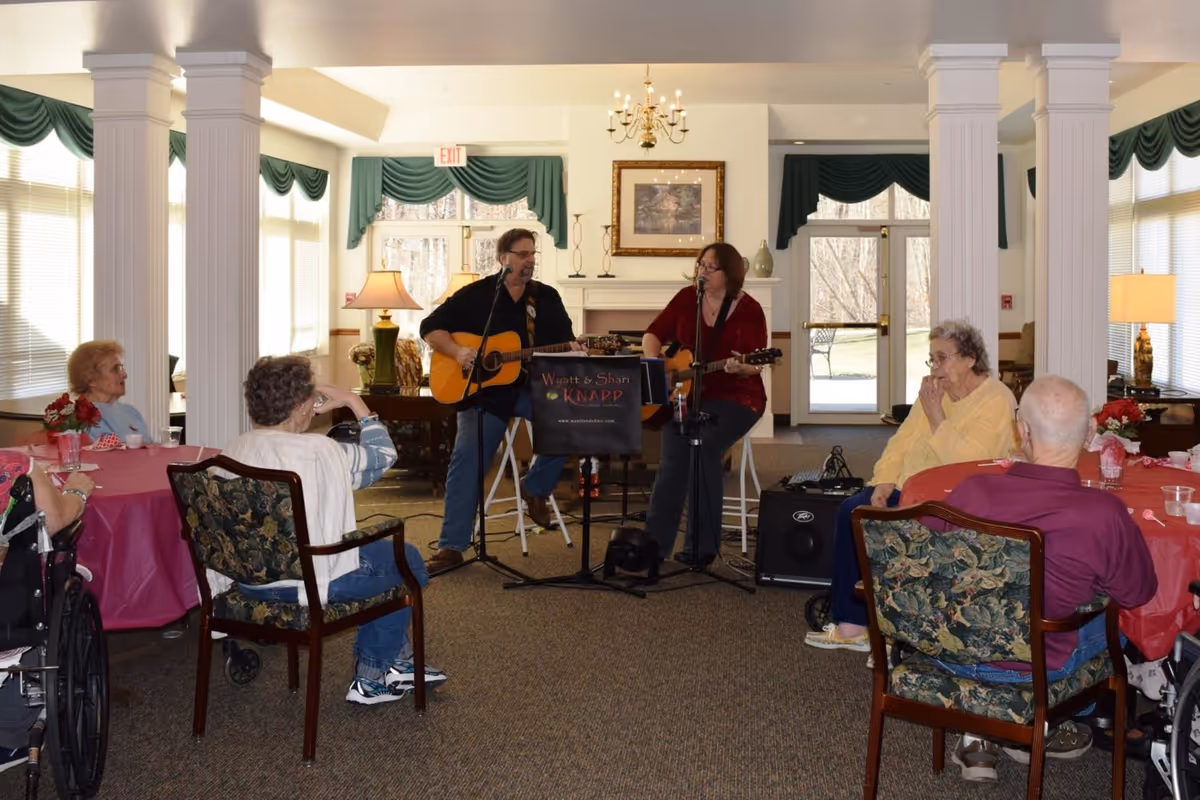 Two musicians playing guitars and singing in a common area of a senior living facility while elderly residents sit around tables covered with red tablecloths, listening. The room has large windows with green valances, white columns, and a chandelier.