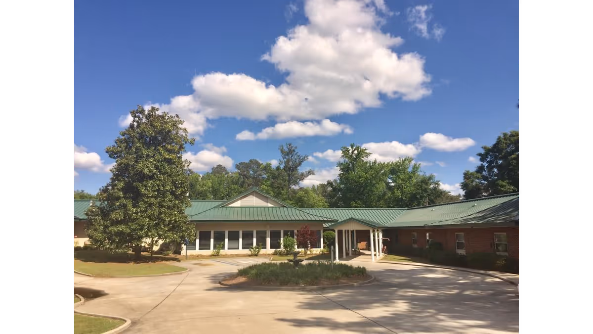 Exterior view of a single-story building with a green metal roof, surrounded by trees and a circular driveway under a partly cloudy blue sky.