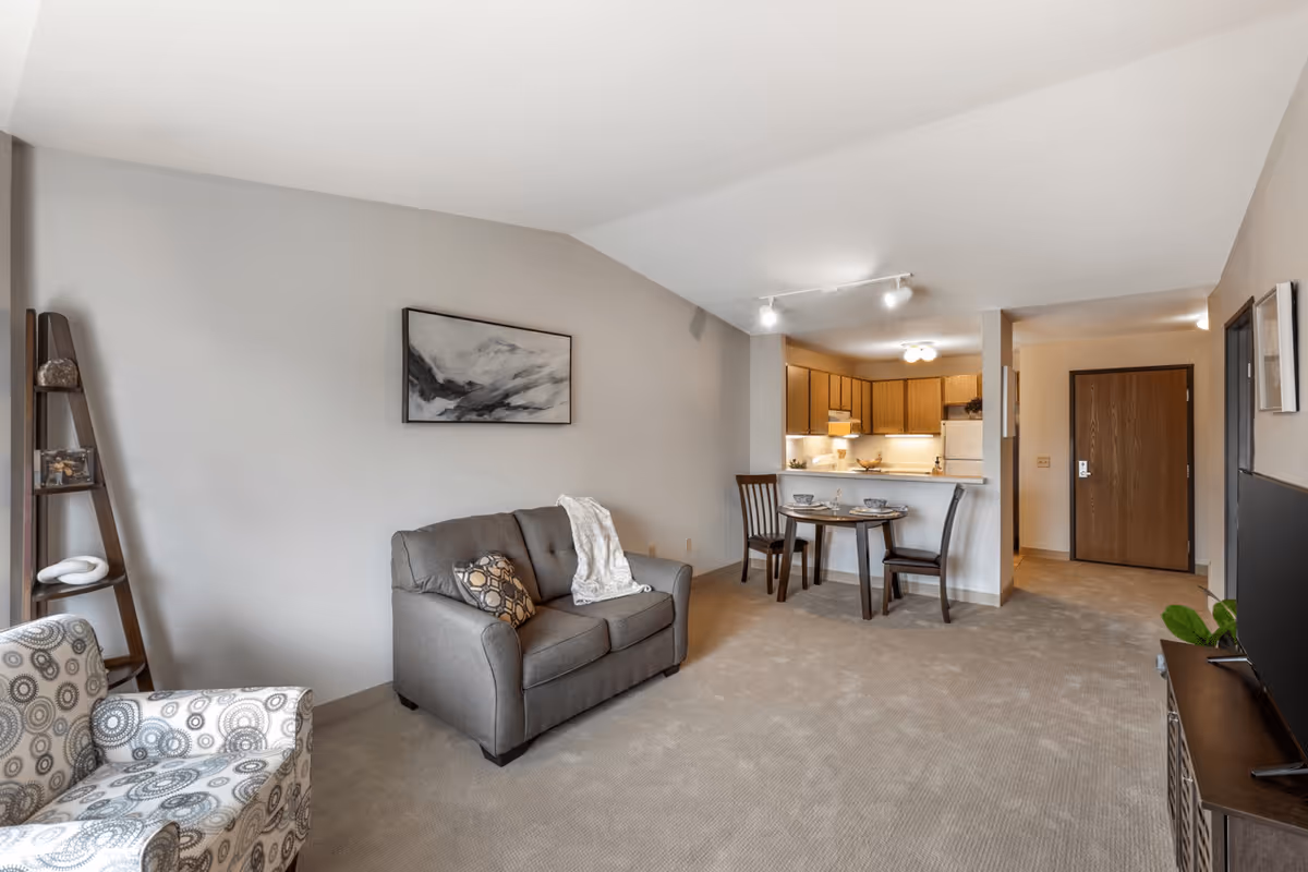 A cozy senior living apartment interior featuring a gray loveseat with a patterned pillow and a white throw blanket, a patterned armchair, a small round dining table set for two, a kitchen with wooden cabinets and white appliances, and a TV on a stand. The walls are light gray and the carpet is beige.