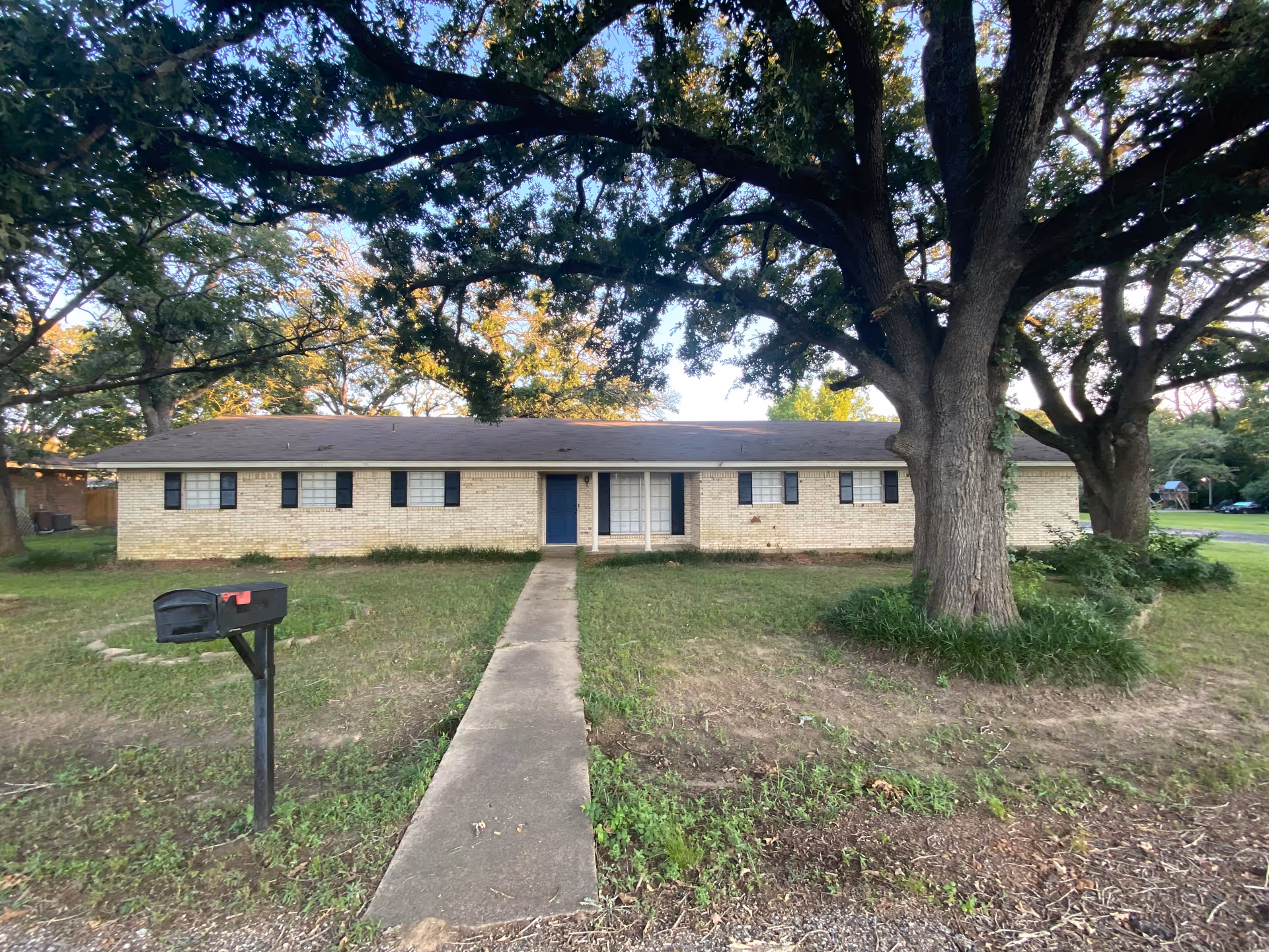 Single-story brick building with a dark shingled roof, multiple windows with black shutters, a blue front door, and a concrete walkway leading to the entrance. Large trees with green foliage are in the front yard, along with a black mailbox on a post near the sidewalk.