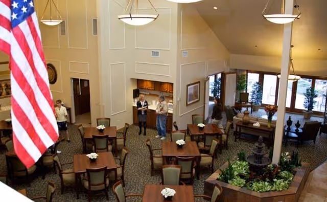 Interior view of a senior living facility dining area with multiple wooden tables and chairs arranged neatly. There are a few people standing and conversing near a kitchenette area. The room features large windows allowing natural light, a small indoor garden with a fountain, and an American flag hanging prominently.