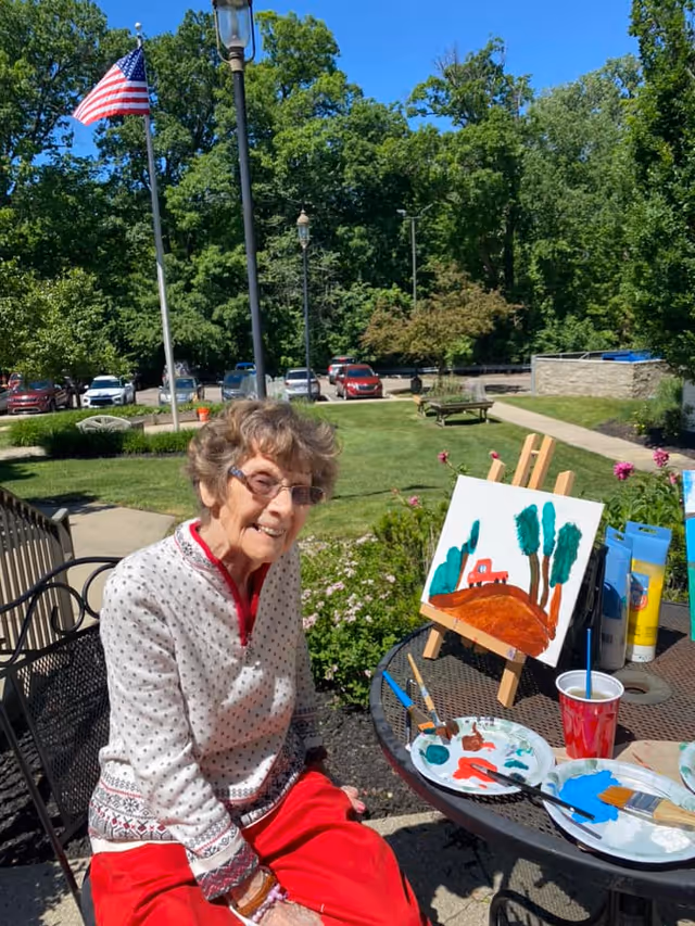 An elderly woman with glasses and curly hair sits outdoors at a metal table with painting supplies. She is smiling and wearing a white sweater with a red pattern and red pants. On the table is a small canvas on an easel with a painting of trees and a red vehicle on a brown hill. The background shows a green lawn, trees, parked cars, and an American flag on a pole.