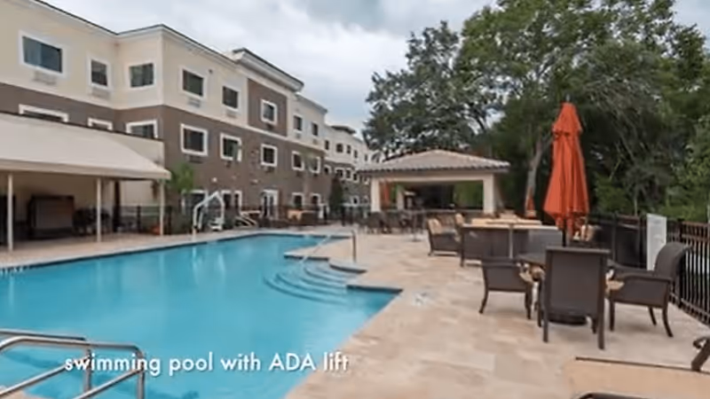 Outdoor swimming pool area at Discovery Village At Deerwood Assisted Living & Memory Care, featuring a pool with an ADA lift, surrounded by lounge chairs, tables with umbrellas, and a covered seating area, with a multi-story building and trees in the background.