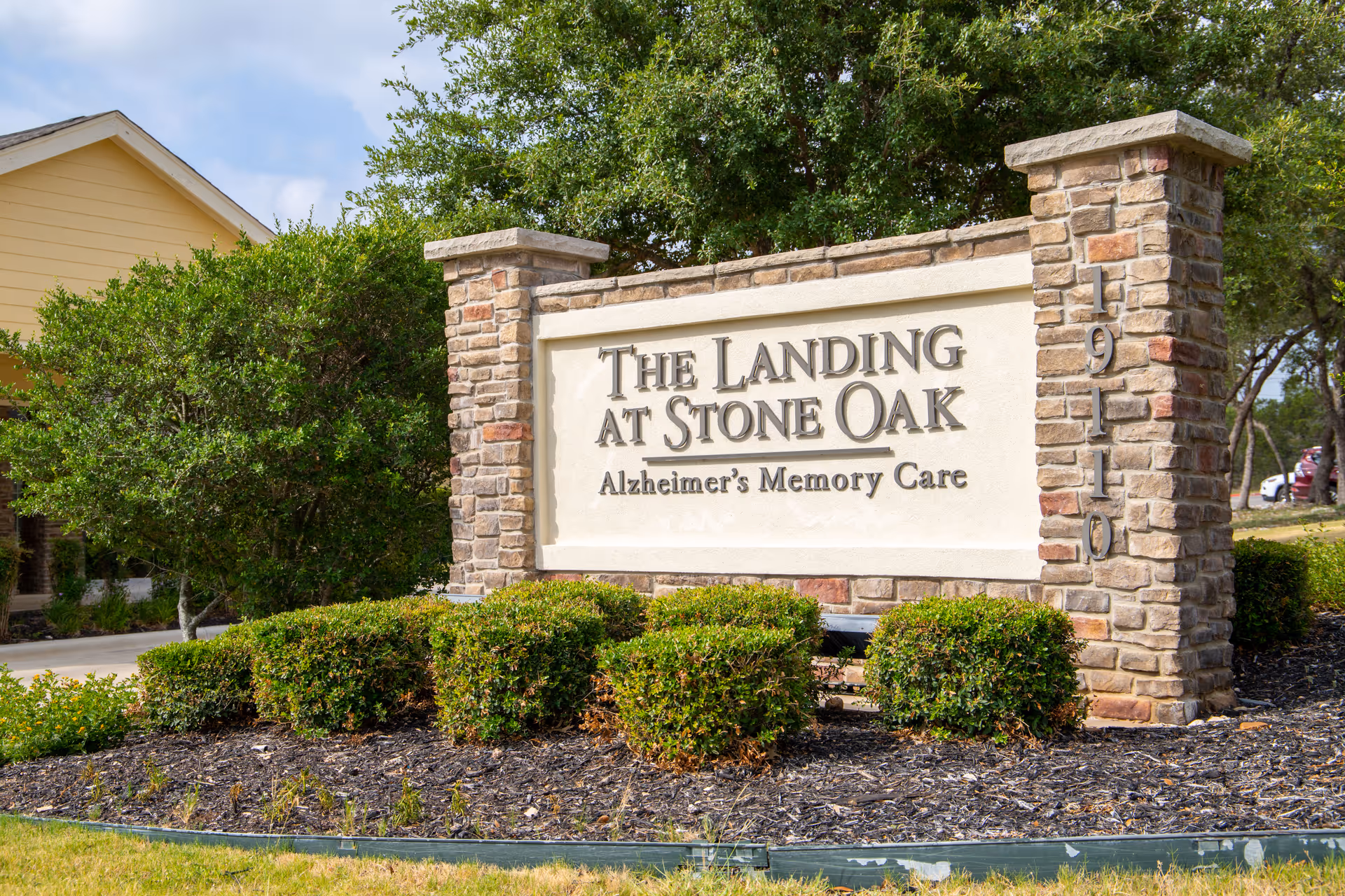 Stone sign for The Landing at Stone Oak Alzheimer’s Memory Care facility surrounded by greenery and bushes, with a yellow building partially visible in the background under a partly cloudy sky.