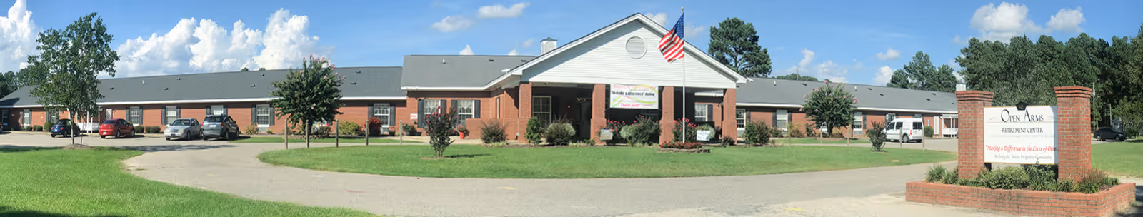 Panoramic view of the front exterior of Open Arms Retirement Center, a single-story brick building with a gray roof, an American flag flying on a flagpole in front, several parked cars, green lawn, and trees under a blue sky with scattered clouds.