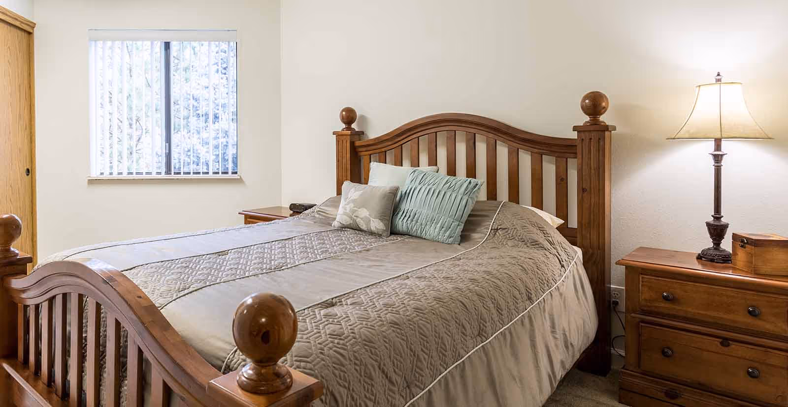 A cozy bedroom with a wooden bed frame featuring rounded finials, a beige quilted bedspread, and three decorative pillows. There is a wooden nightstand with a lamp and a small wooden box on the right side of the bed. A window with vertical blinds is on the left wall, allowing natural light into the room.
