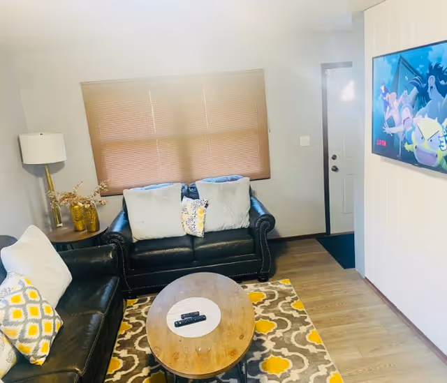 A cozy living room with two black leather sofas adorned with white and yellow patterned cushions. A wooden oval coffee table with two remote controls sits on a patterned rug with yellow and gray designs. A floor lamp and decorative vases are on a side table next to one sofa. A window with closed blinds is behind one sofa, and a flat-screen TV is mounted on the wall opposite the sofas. The room has light-colored walls and wooden flooring.