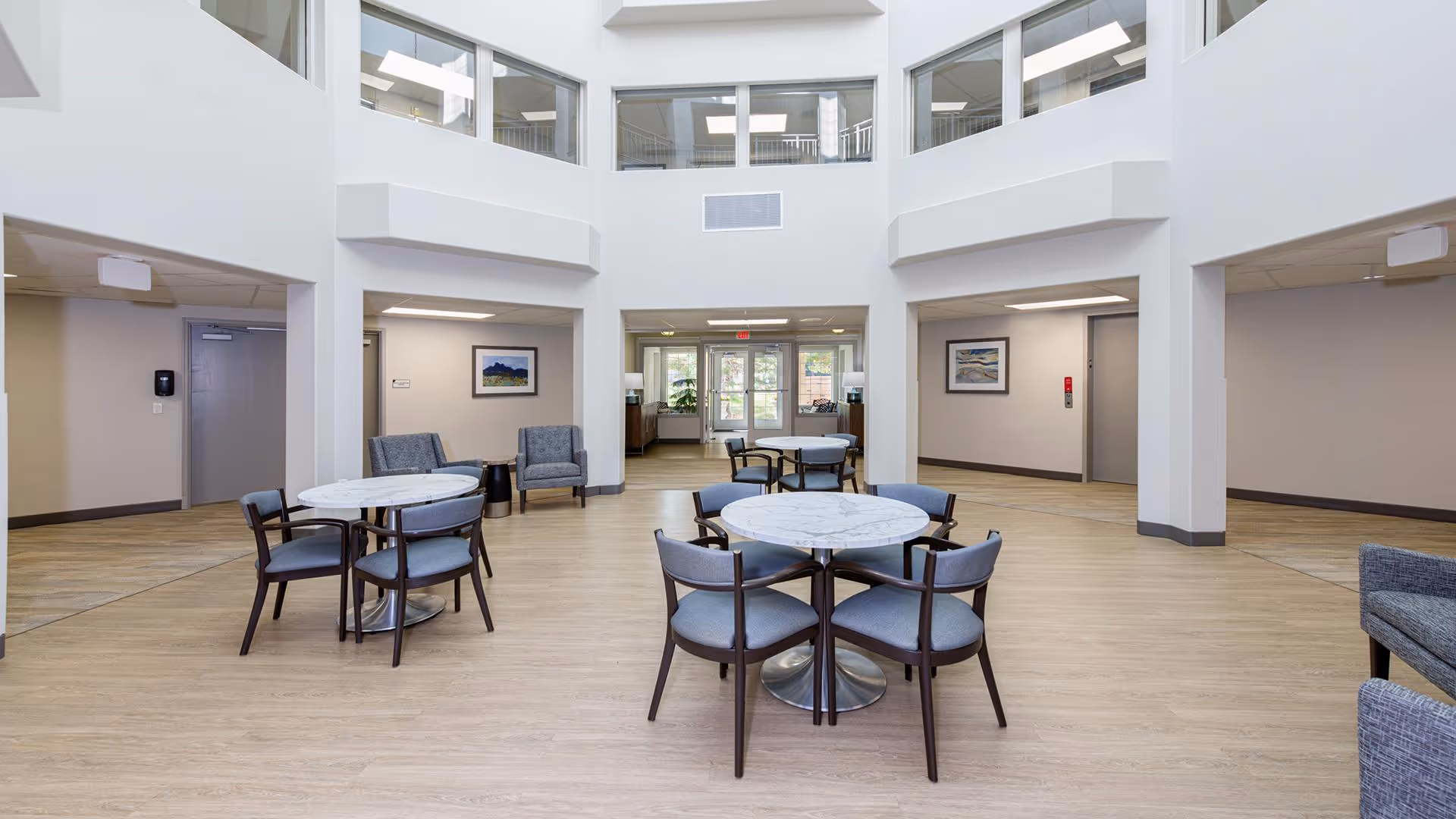 A bright, spacious common area in a senior living facility with round tables and chairs arranged on a light wood floor. There are two armchairs against the wall, framed artwork, and large windows on the upper level letting in natural light. The area has a clean, modern design with white walls and a high ceiling.