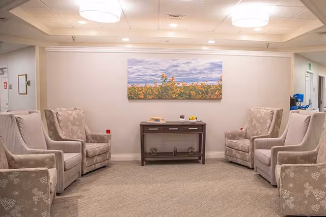 A cozy seating area in a senior living facility with four beige patterned armchairs arranged around a small dark wooden console table. On the table are a few decorative items and books. Above the table hangs a large landscape painting of a field of sunflowers under a partly cloudy sky. The room has soft lighting from ceiling fixtures and neutral-colored walls and carpet.
