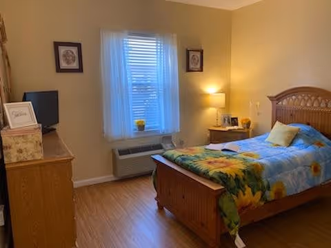 A cozy bedroom with a wooden bed featuring a colorful sunflower-themed blanket and a yellow pillow. There is a wooden nightstand with a lamp and framed photos next to the bed. A window with sheer white curtains is centered on the wall, and a wooden dresser with a small TV and decorative items is on the left side of the room. The floor is wooden, and the walls are painted a warm beige color.