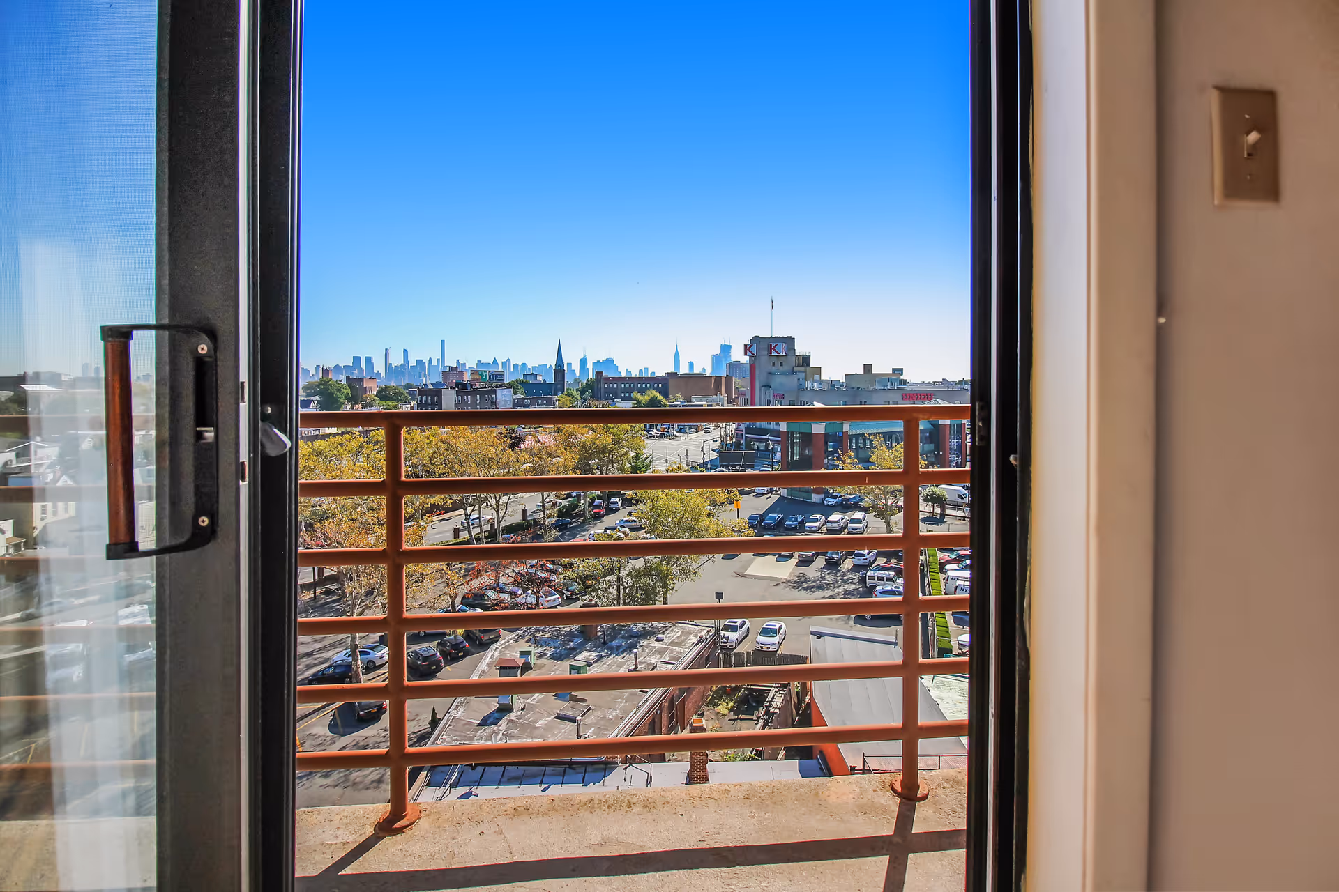 View through a sliding glass door onto a balcony with a metal railing overlooking a cityscape with buildings, trees, and parked cars under a clear blue sky.