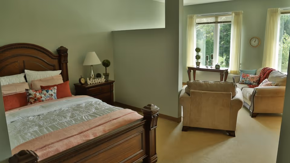 A cozy room with a wooden bed featuring white and coral bedding and multiple decorative pillows. Next to the bed is a wooden nightstand with a lamp, a clock, a small plant, and a decorative sign that says 'blessed'. The room also has a sitting area with a beige armchair and a matching sofa adorned with colorful pillows. Large windows with light curtains let in natural light and offer a view of greenery outside.