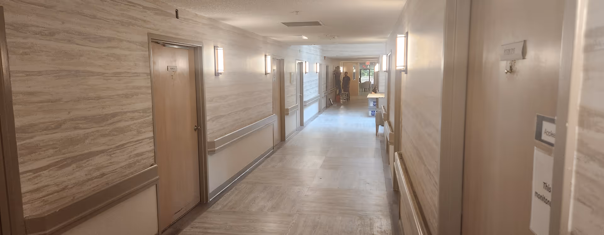 A long, well-lit hallway in an assisted living facility with beige walls and wooden doors on both sides. Handrails run along the walls, and a person is visible at the far end of the corridor near an exit door with glass panels.