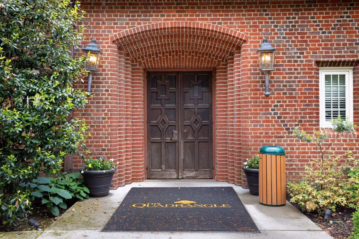 Entrance to a brick building with a double wooden door under a brick archway. There are two lit lantern-style wall lights on either side of the door, potted plants, a trash can, and a black mat with the text 'The Quadrangle' in yellow in front of the door.