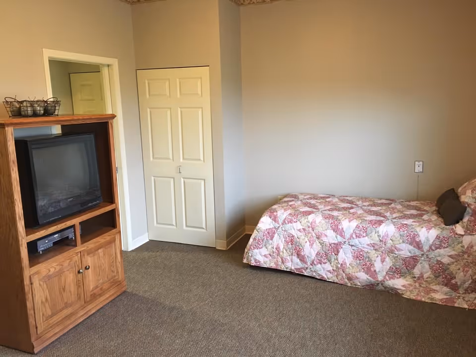 A simple bedroom with a single bed covered in a pink and white patterned quilt, a wooden TV stand with an older-style television and a DVD player, a closed closet with double doors, and beige walls and carpet.