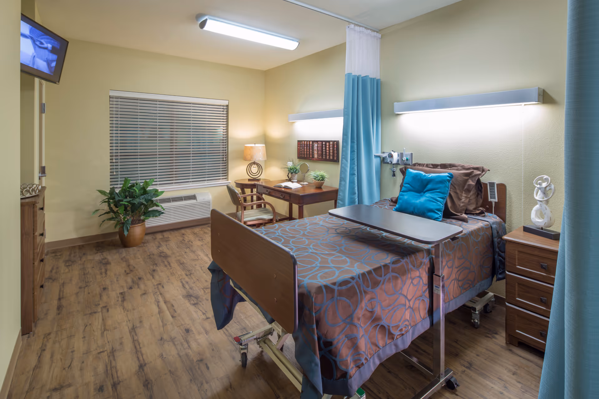 A well-lit senior living facility bedroom with a hospital-style bed covered in a brown and blue patterned blanket, a blue pillow, and an overbed table. The room has wooden flooring, a small wooden desk with a chair, a potted plant near the window with blinds, a bedside table with a decorative sculpture, and a wall-mounted TV in the corner.