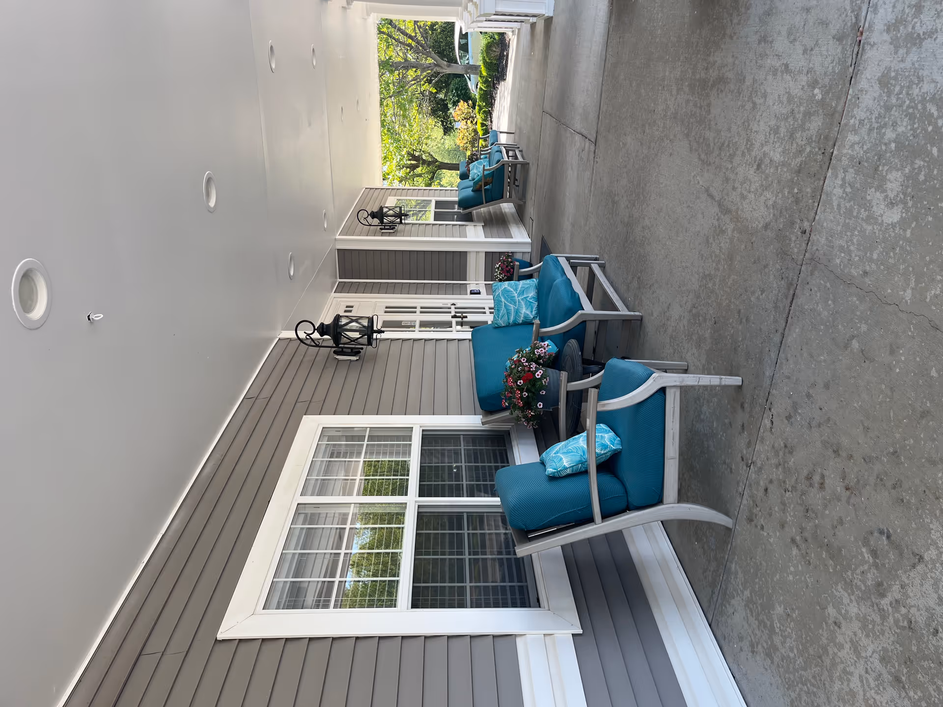 Covered outdoor porch area with gray siding walls, white trim windows, and multiple blue cushioned chairs with patterned pillows. Small round tables with flower pots are placed between the chairs. Black lantern-style wall lights are mounted on the wall. Trees and greenery are visible in the background.