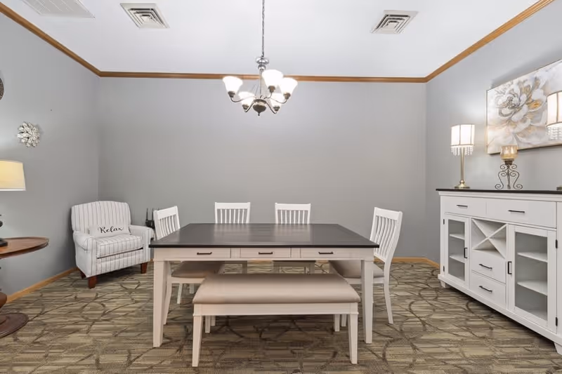A dining room with a rectangular dark wood table surrounded by four white chairs and a cushioned bench. To the left, there is a white armchair with a 'Relax' pillow. On the right side, there is a white sideboard with drawers and glass doors, topped with two lamps and a decorative candle holder. The walls are painted light gray with wooden trim, and a chandelier hangs above the table.