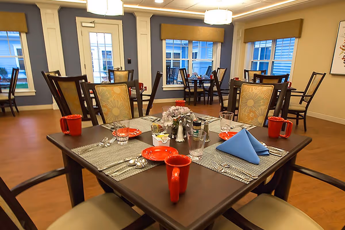 A dining room in a senior living facility with several tables and chairs. The tables are set with red mugs, red plates, silverware, glasses, and a centerpiece with flowers. The room has large windows with beige valances and blue walls, and the floor is wooden.