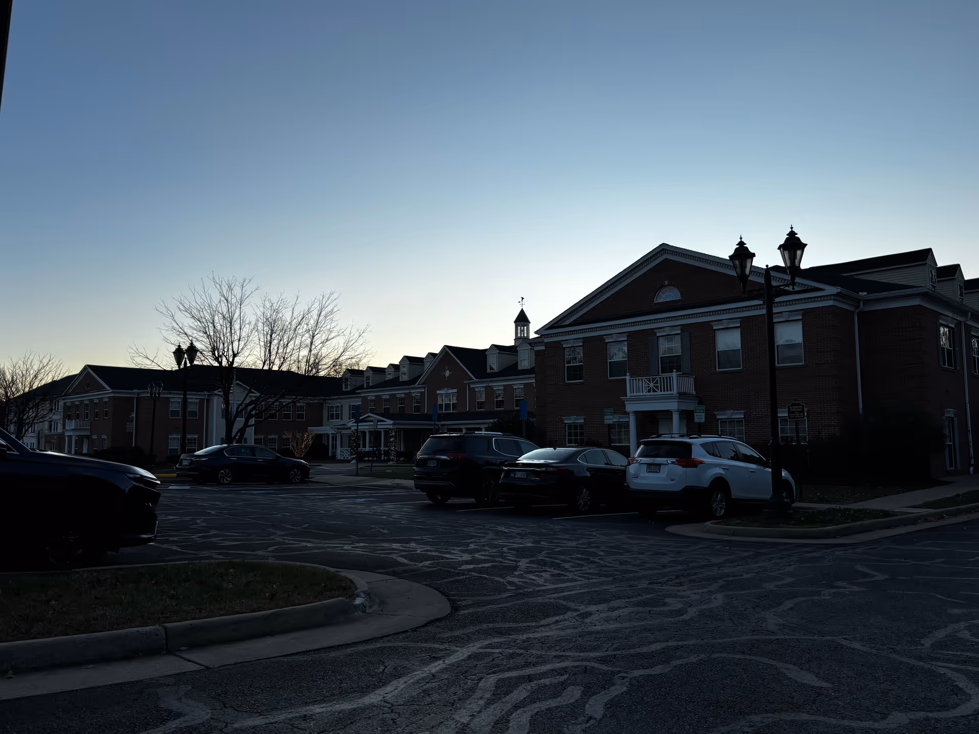 Exterior view of a senior living facility building during dusk with several parked cars in front and bare trees around the parking lot.