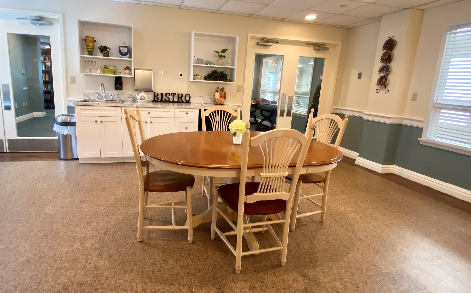 A dining area with a round wooden table and four chairs. Behind the table is a white cabinet with a sink, shelves with decorative items, and a sign that reads 'BISTRO'. There are double glass doors and windows with blinds on the right side, and the walls are painted in a two-tone style with white on top and green on the bottom.