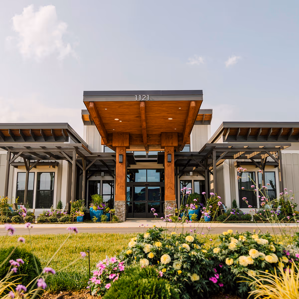 Front exterior view of a modern senior living facility building with a wooden canopy entrance, large windows, and landscaped flower beds with colorful flowers and greenery in the foreground under a partly cloudy sky.