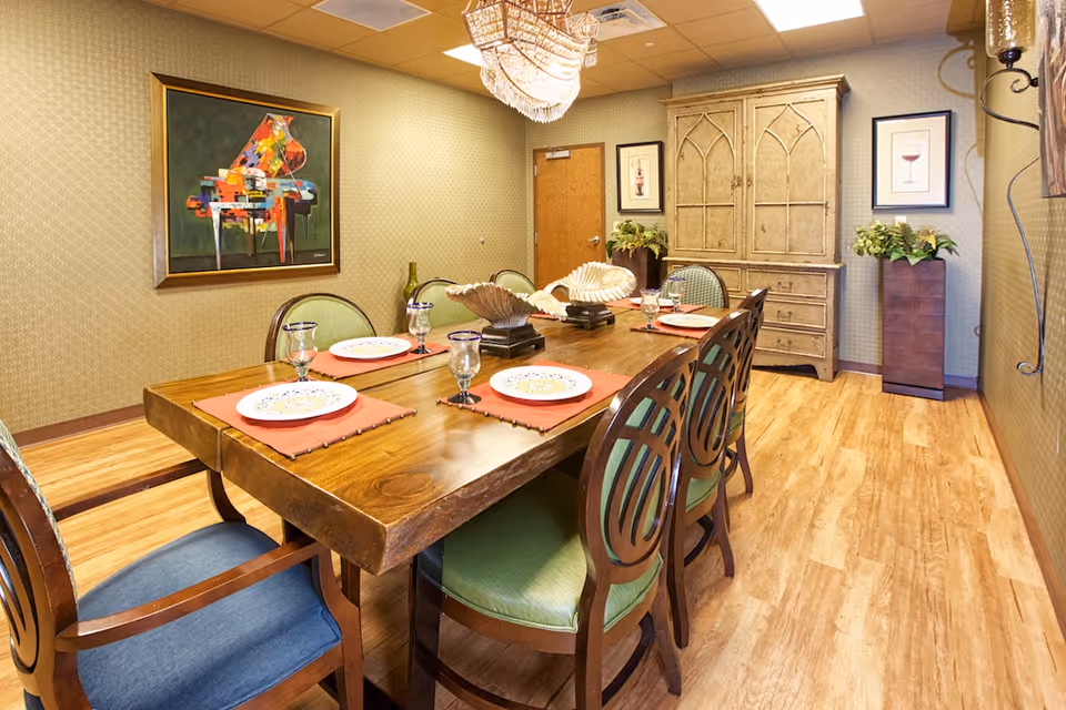 A dining room with a wooden table set for six people. The table has orange placemats, decorative plates, and glassware. There are six chairs with green and blue cushions around the table. The room has wooden flooring, patterned wallpaper, a chandelier, a large decorative cabinet, two framed pictures on the wall, and two tall plant stands with greenery.