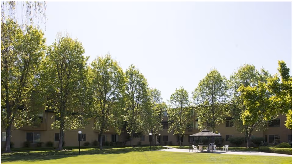 Outdoor view of Bethel Retirement Community showing a grassy lawn with several trees and a paved area with a gazebo and chairs. The building is visible in the background under a clear sky.