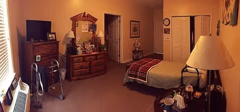 A cozy bedroom with a bed covered in a plaid blanket, a wooden dresser with a mirror and various personal items, a television on a chest of drawers, a walker, a lamp on a bedside table, and a closet with double doors.