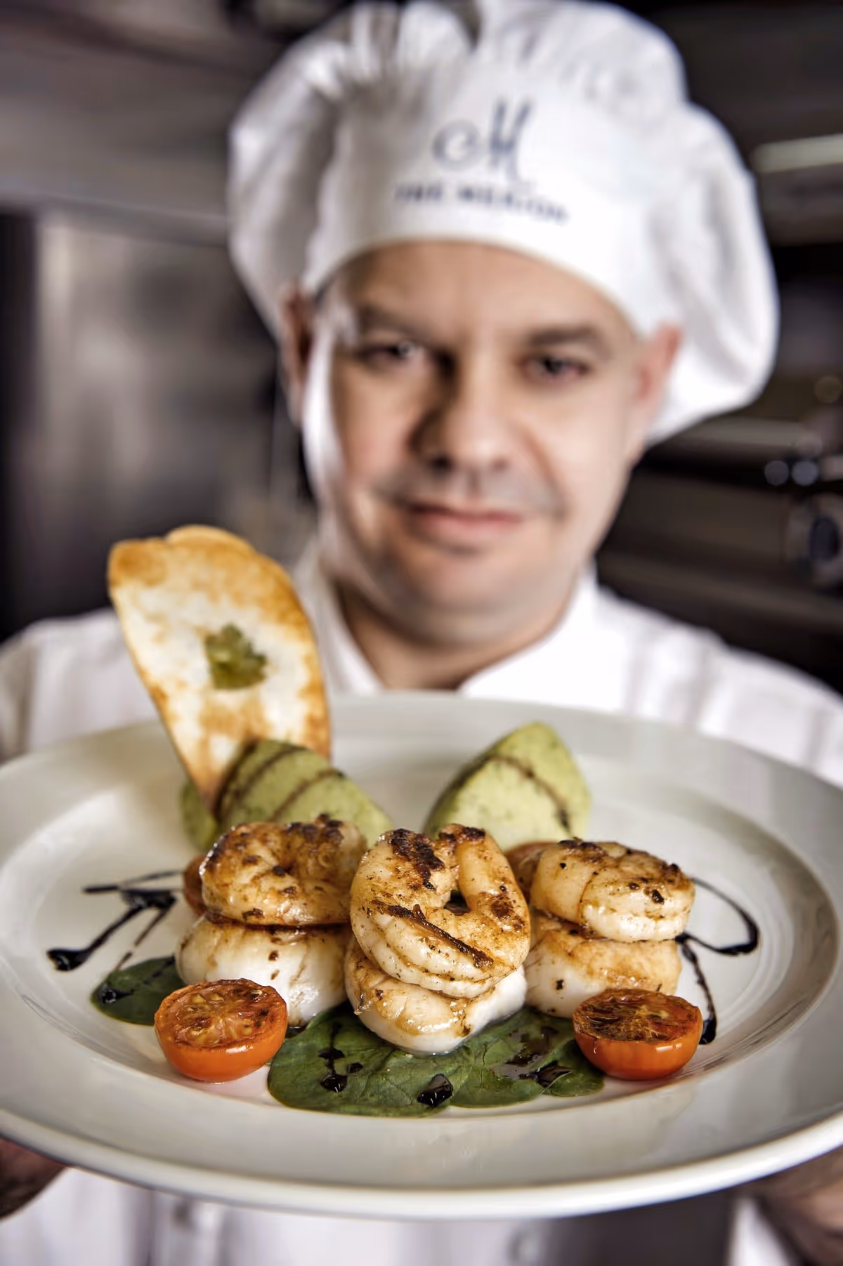 A chef wearing a white hat and uniform is holding a plate of grilled shrimp served with cherry tomatoes, green garnish, and a toasted bread slice, presented in a professional kitchen setting.
