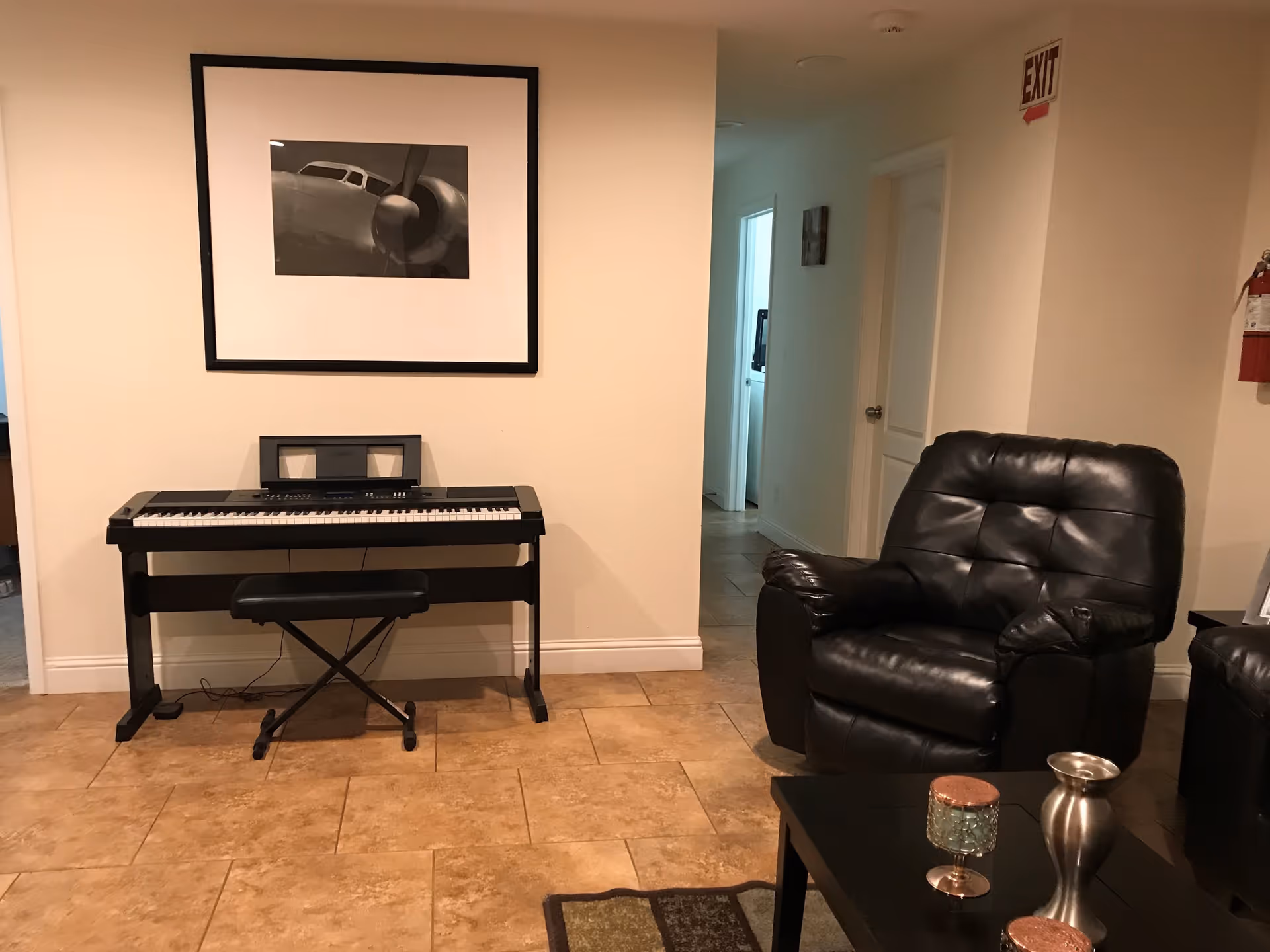 Interior view of a room with a black electronic keyboard on a stand and a matching bench against a beige wall. Above the keyboard is a framed black and white picture of an airplane propeller. To the right, there is a black leather recliner chair next to a dark wooden coffee table with decorative items. The floor is tiled, and a hallway with doors is visible in the background. An exit sign and a fire extinguisher are mounted on the wall.