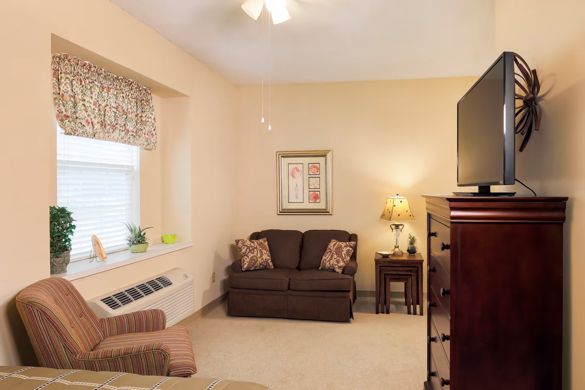 A cozy living room area with a brown loveseat adorned with two patterned pillows, a wooden side table with a lamp and small plant, a striped armchair, a window with floral valance and blinds, and a wooden dresser with a flat-screen TV on top.
