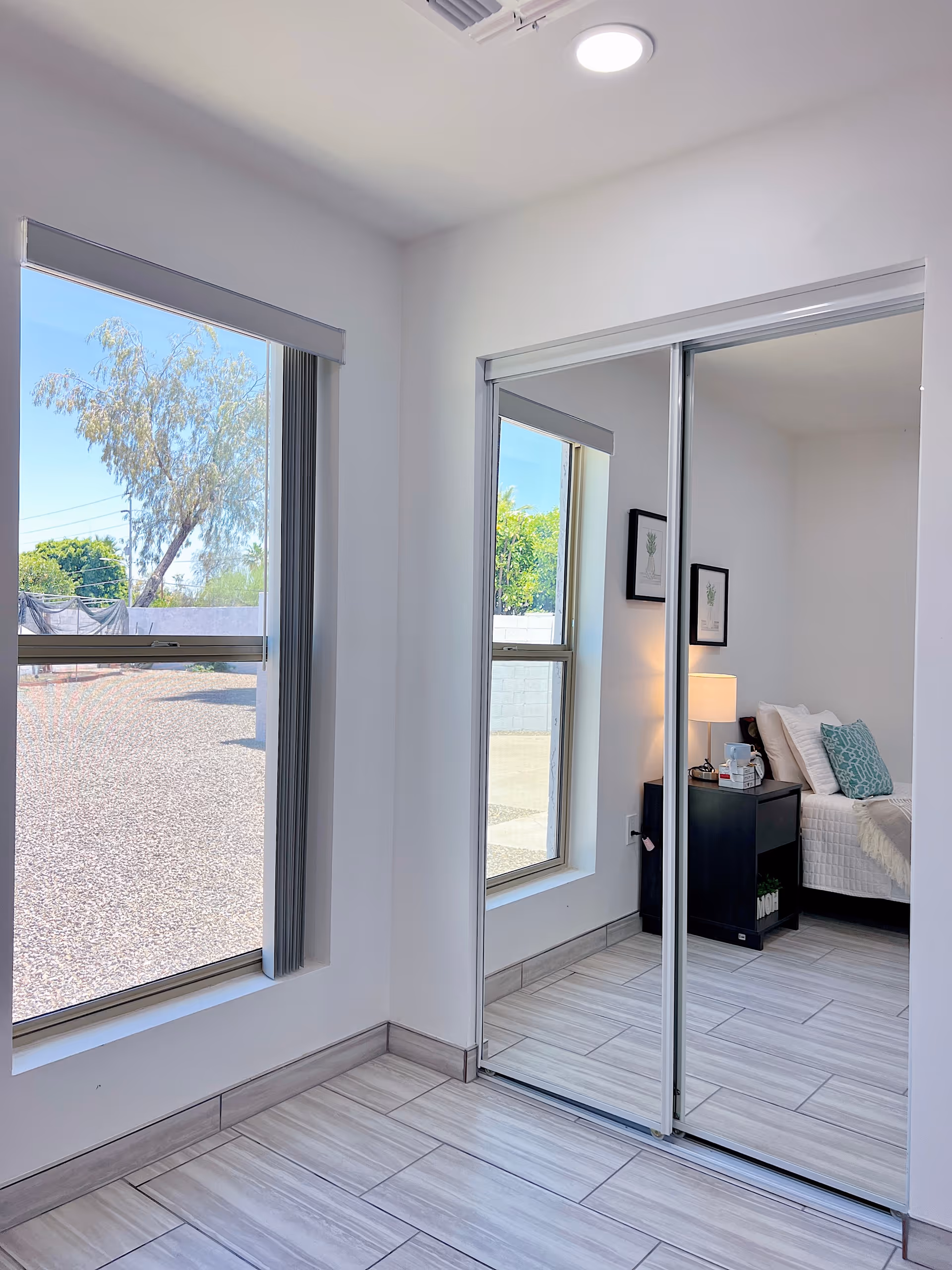 A bright room with large windows showing an outdoor view of trees and gravel. The room has light-colored tiled flooring and white walls. A mirrored sliding closet door reflects a bedside table with a lamp, some framed artwork on the wall, and a bed with pillows and a blanket.