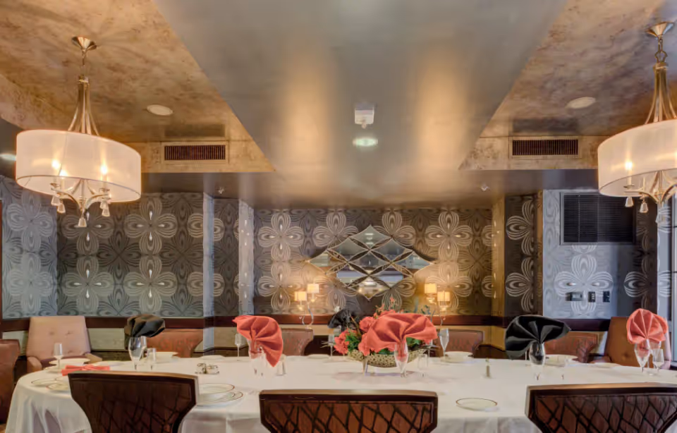 Elegant dining room with a large oval table covered with a white tablecloth, set with plates, glasses, and folded napkins in black and red. The room features patterned wallpaper, two chandeliers, cushioned chairs, and a decorative mirror on the wall.