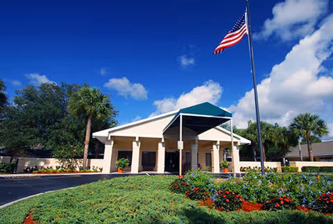 Front entrance of a senior living facility with a covered porte-cochere, landscaped flower beds, palm trees and an American flag under a blue sky.