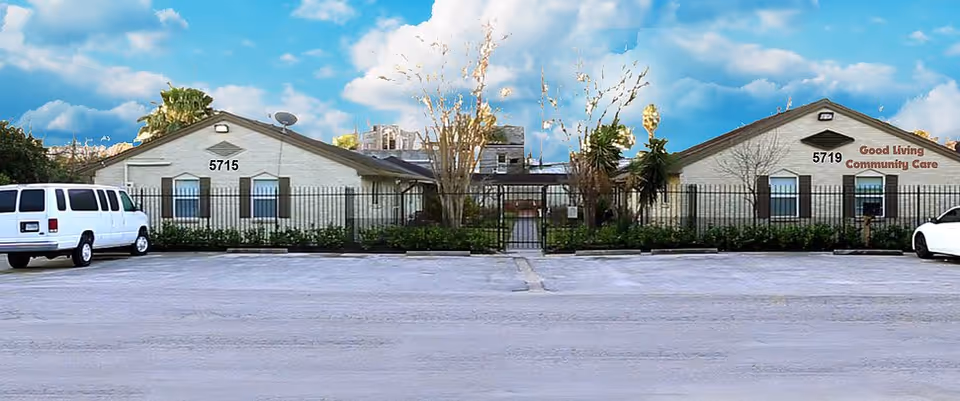 Exterior view of Good Living Community Care facility showing two single-story buildings with beige walls and dark window shutters, separated by a gated walkway. There are two parked vehicles, a white van on the left and a white car on the right, with a clear blue sky and some trees in the background.